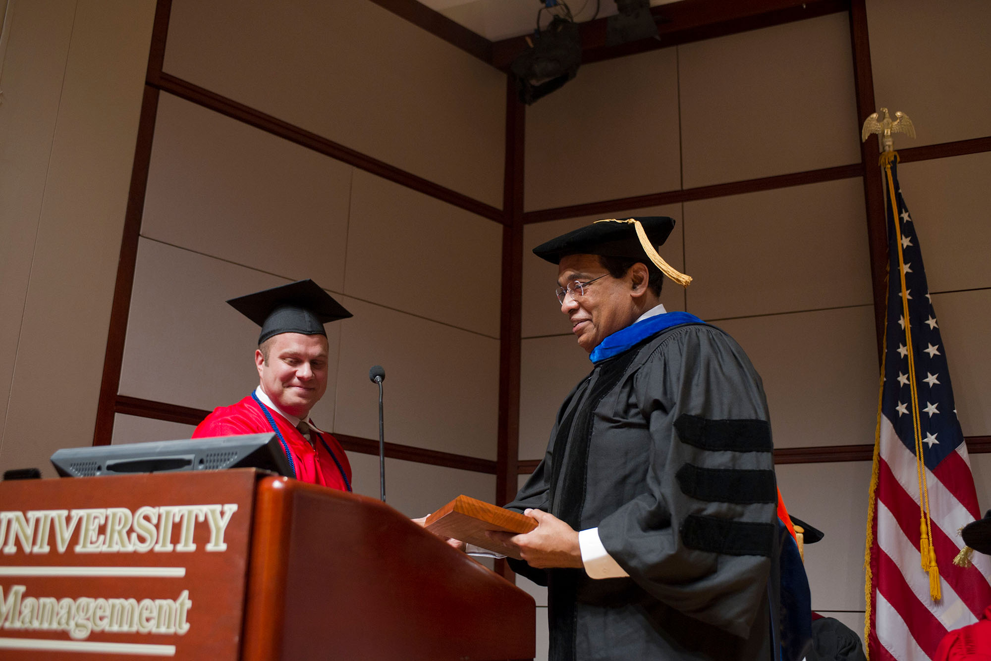 Photo: Two men on stage. The one on the left is wearing a red gown and cap and the man on the right is wearing a black gown and cap