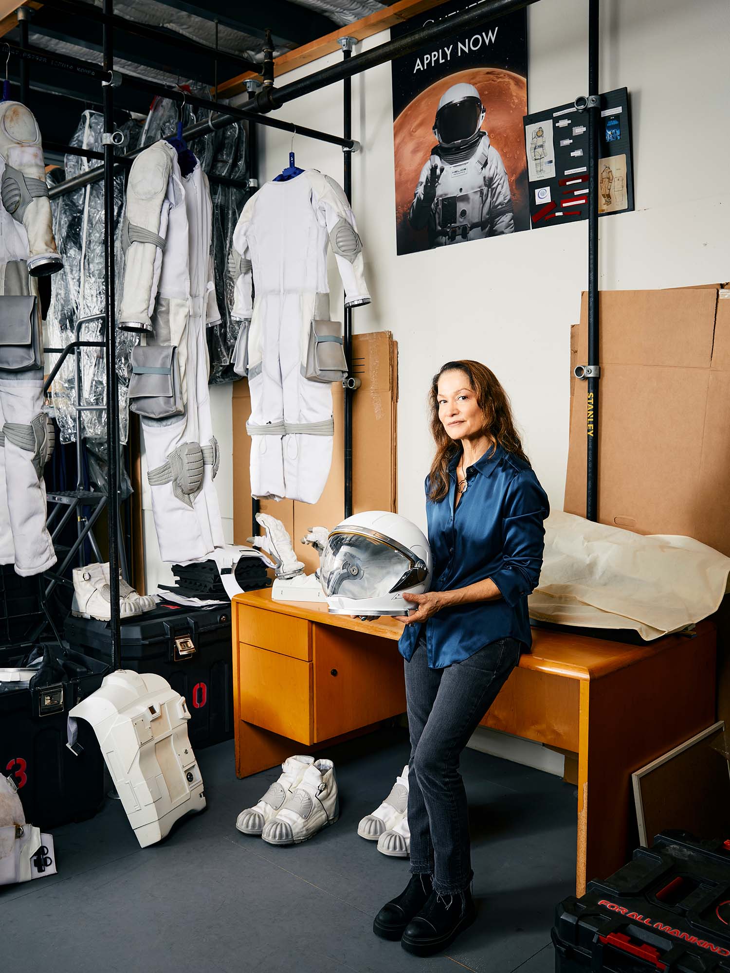 Photo: A woman leaning against a desk as she holds an astronaut helmet. The wall behind her has astronaut suits across it and a poster with an astronaut that says "Apply Now"