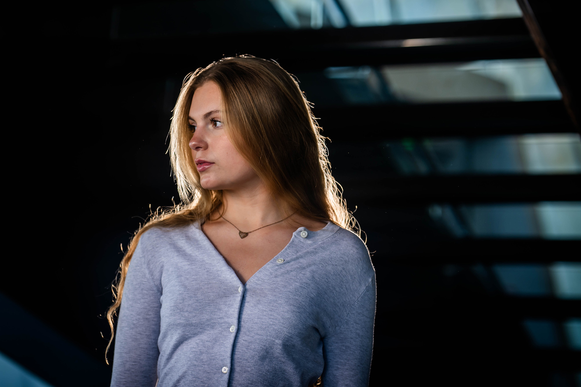 Photo: A blonde female student wearing a gray cardigan and looking off to the side as she stands under a staircase