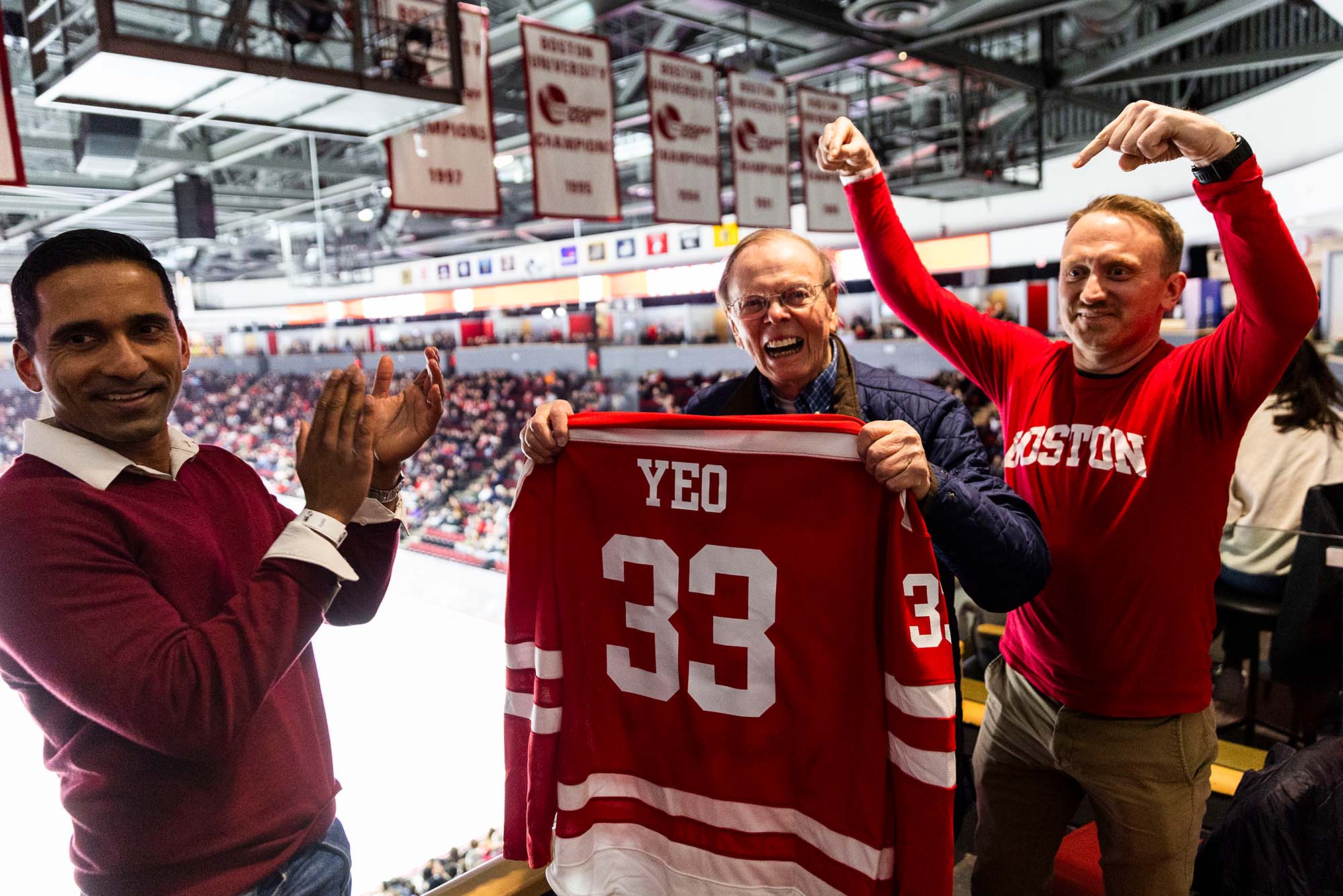 Photo: A 91-year-old man holding up a BU Hockey jersey with his name on it as two men next to him celebrate