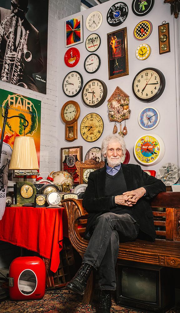 Photo: A distinguished man with white hair sits with hands folded in front of a wall of ornate clocks and other items