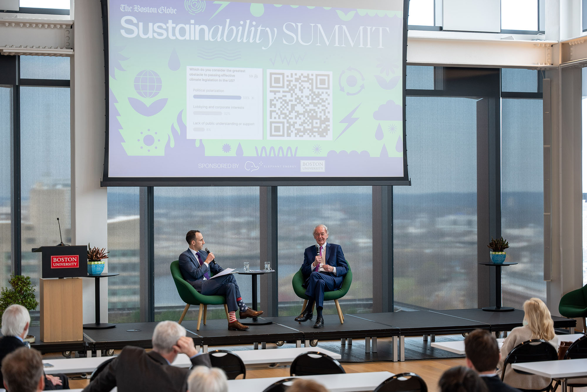 Photo: Two men sit on a stage with a projector above them with a screen that reads SUSTAINABILITY SUMMIT