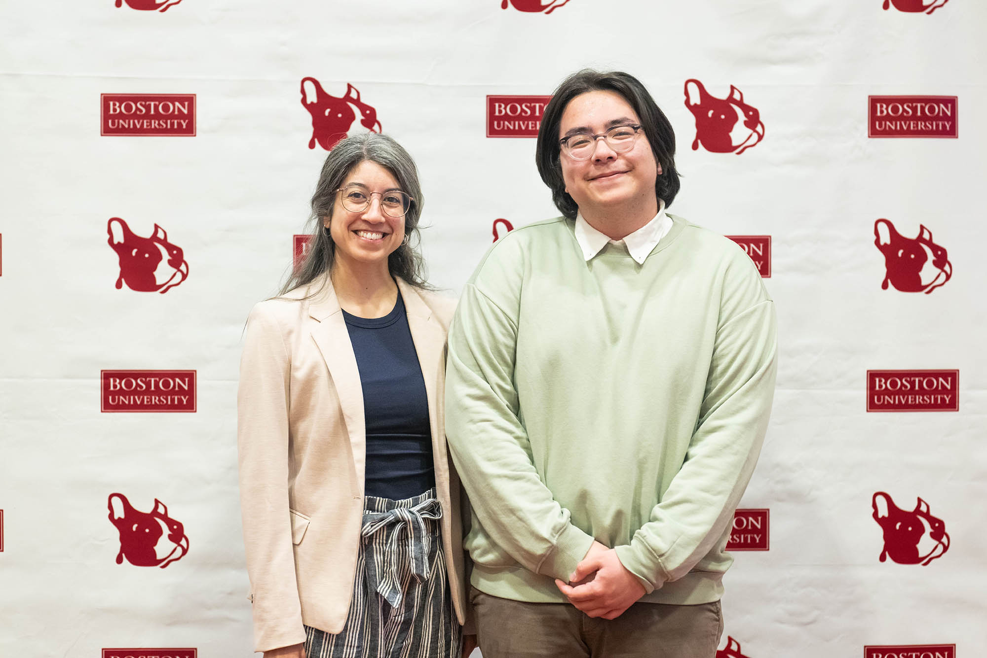 Photo: An older woman with a pink blazer smiles next to a younger man with chin length black hair.