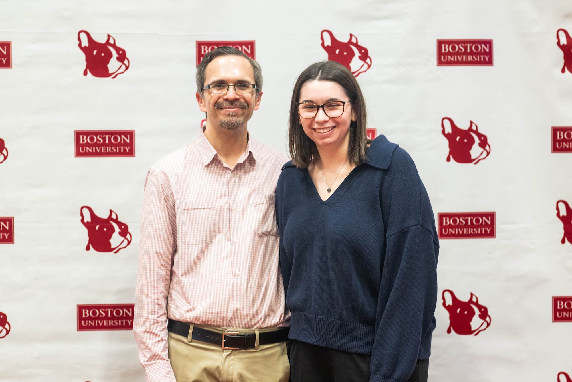 Photo: A older man with a pink shirt poses with a young woman with a fray zipup for a portrait photo.