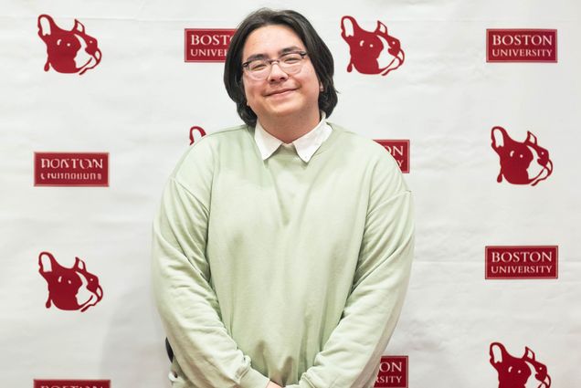 Photo: A young man with chin length black hair poses for a photo in a green sweater.