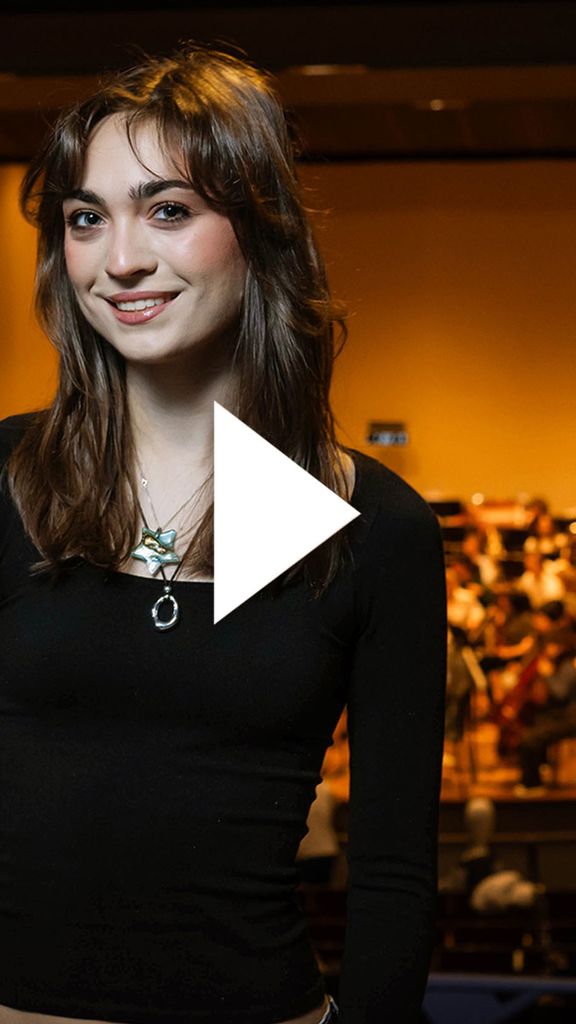 Photo: A girl with brown hair and bangs wearing a long sleeve black shirt and smiling in front of a stage. There is a video play button over the image