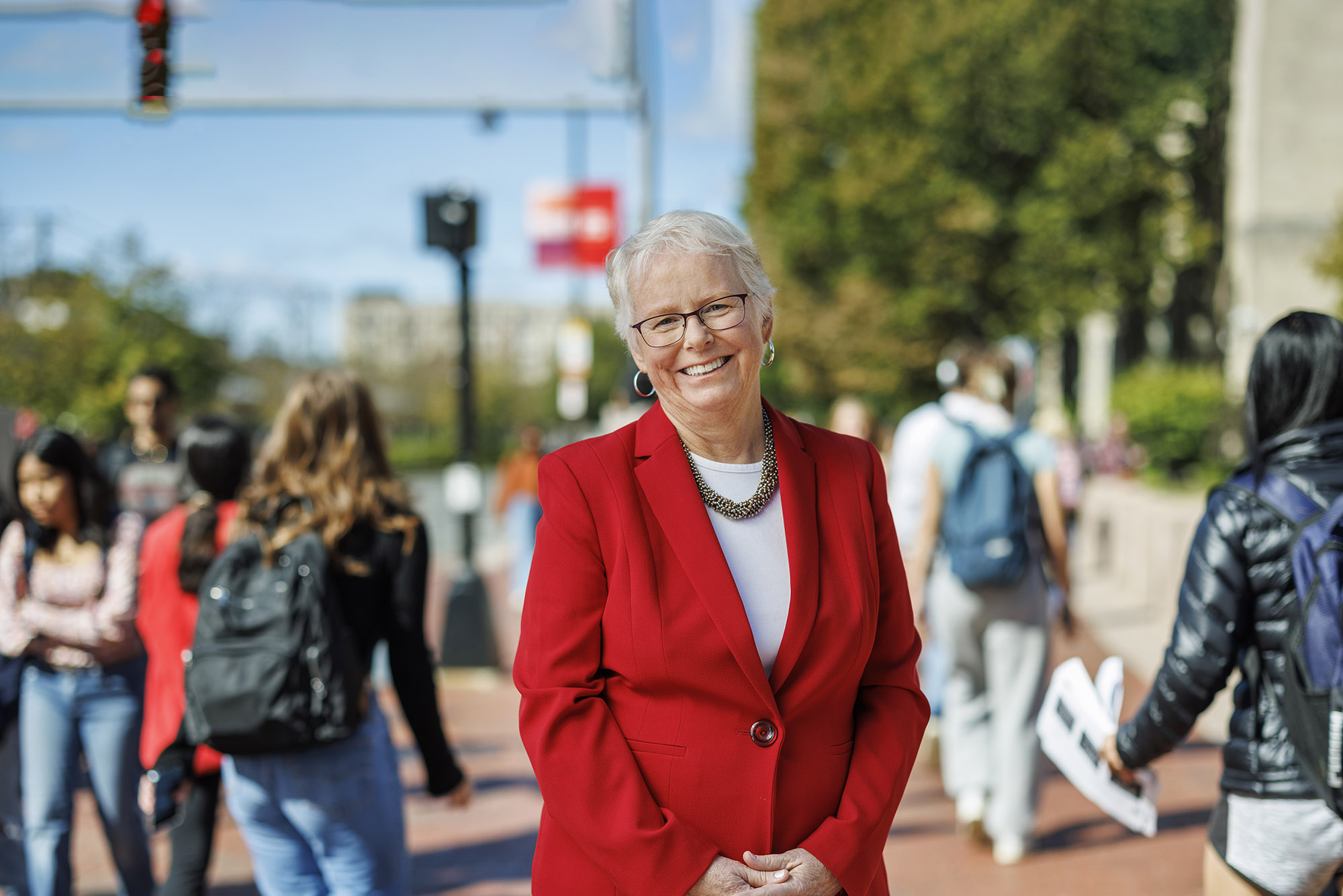 Photo: Portrait of Boston University Provost Jean Morrison on Commonwealth Ave. Asmiling white woman with short, silver white hair and wearing glasses, a white blouse, and red blazer poses with fingers clasped in front of her on a bustling sidewalk. Background and passerby are blurry.