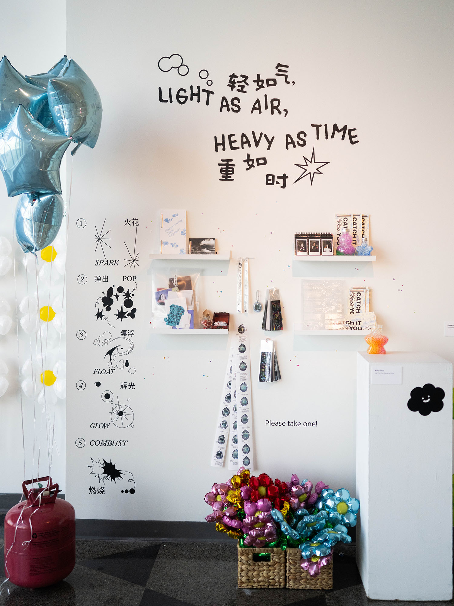 Photo: An art exhibit featuring balloons floating and a basket full of ones that can be taken by viewers. Writing on the wall says "Light as air, heavy as time"
