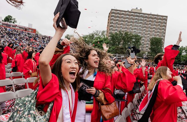 Photo: A group of 波士顿大学 graduates in red gowns and caps celebrate at an outdoor graduation ceremony. Some are raising their hands and holding diplomas, while others have their faces blurred. The background shows a large crowd of spectators in a stadium and a tall building.