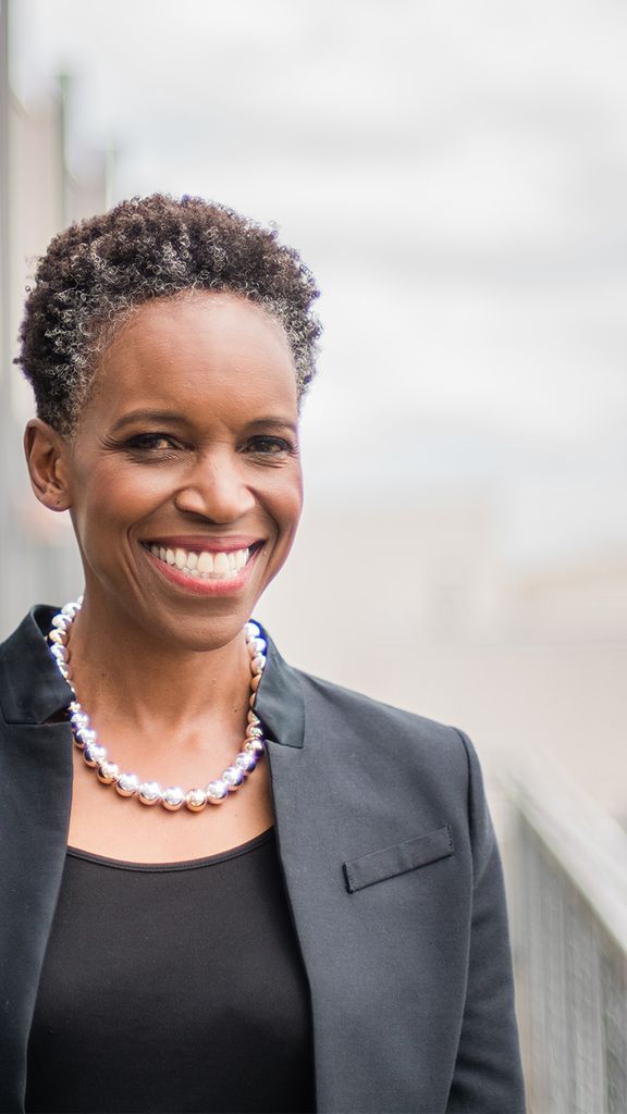 Photo: A black woman with short hair wearing a black blazer over a black shirt with a pearl necklace, smiling for a portrait in front of a blurred out cityscape
