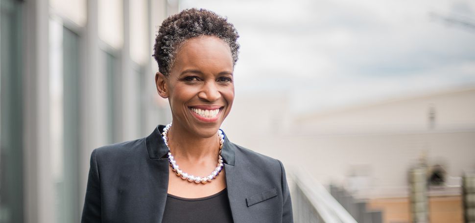 Photo: A black woman with short hair wearing a black blazer over a black shirt with a pearl necklace, smiling for a portrait in front of a blurred out cityscape