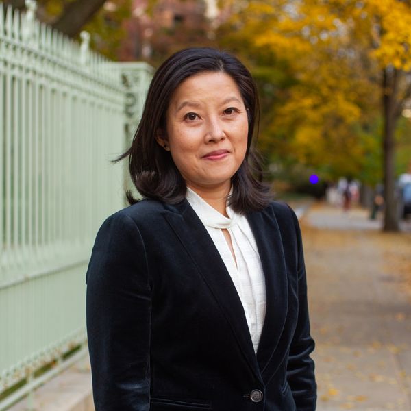 Dr Alice Tseng wearing a dark suit and white shirt. She is standing outside, in the foreground of of an autumnal, tree-lined, wide sidewalk.
