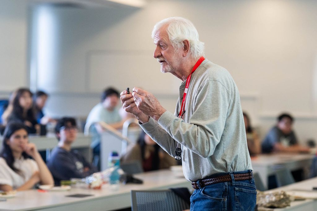 Photo: An older man in jeans and a buttoned shirt holds a rock as he teaches a class.