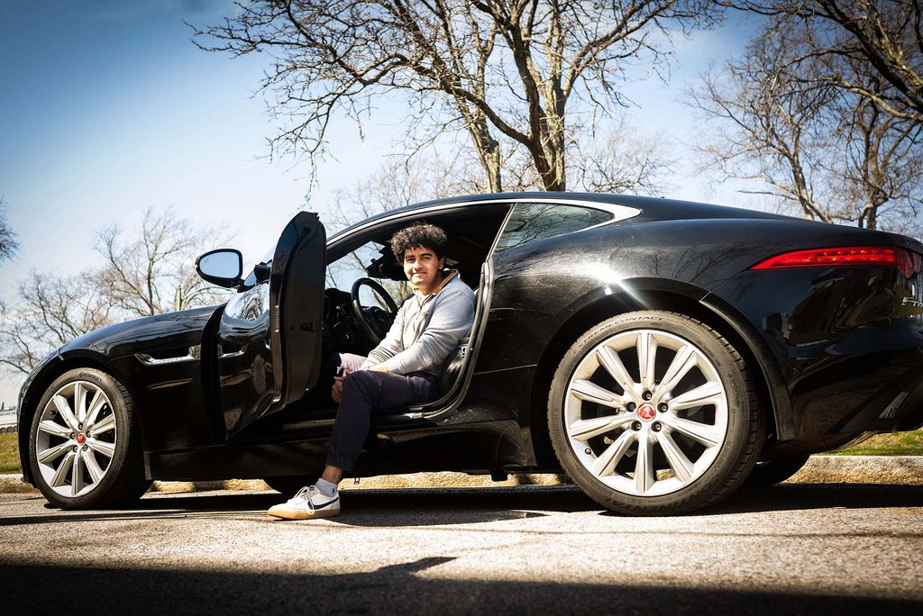 Photo: A male BU student sitting in a black sedan with the door open and one leg on the sidewalk outside of the car.