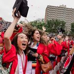 Photo: A group of Boston University graduates in red gowns and caps celebrate at an outdoor graduation ceremony. Some are raising their hands and holding diplomas, while others have their faces blurred. The background shows a large crowd of spectators in a stadium and a tall building.
