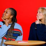 Photo: Two female BU students wearing blue sweaters posing inquisitively in front of a red background as they sit behind a table