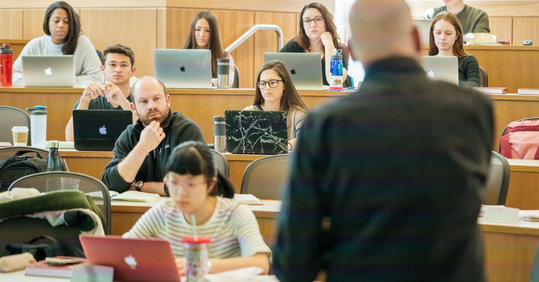Law professor with his back to the camera teaching students gathered before him in a lecture hall listening intently.