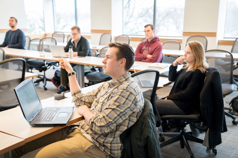 handful of law students seated in a lecture hall with laptops, male in beige flannel in the front answering a prompt from a professor off screen