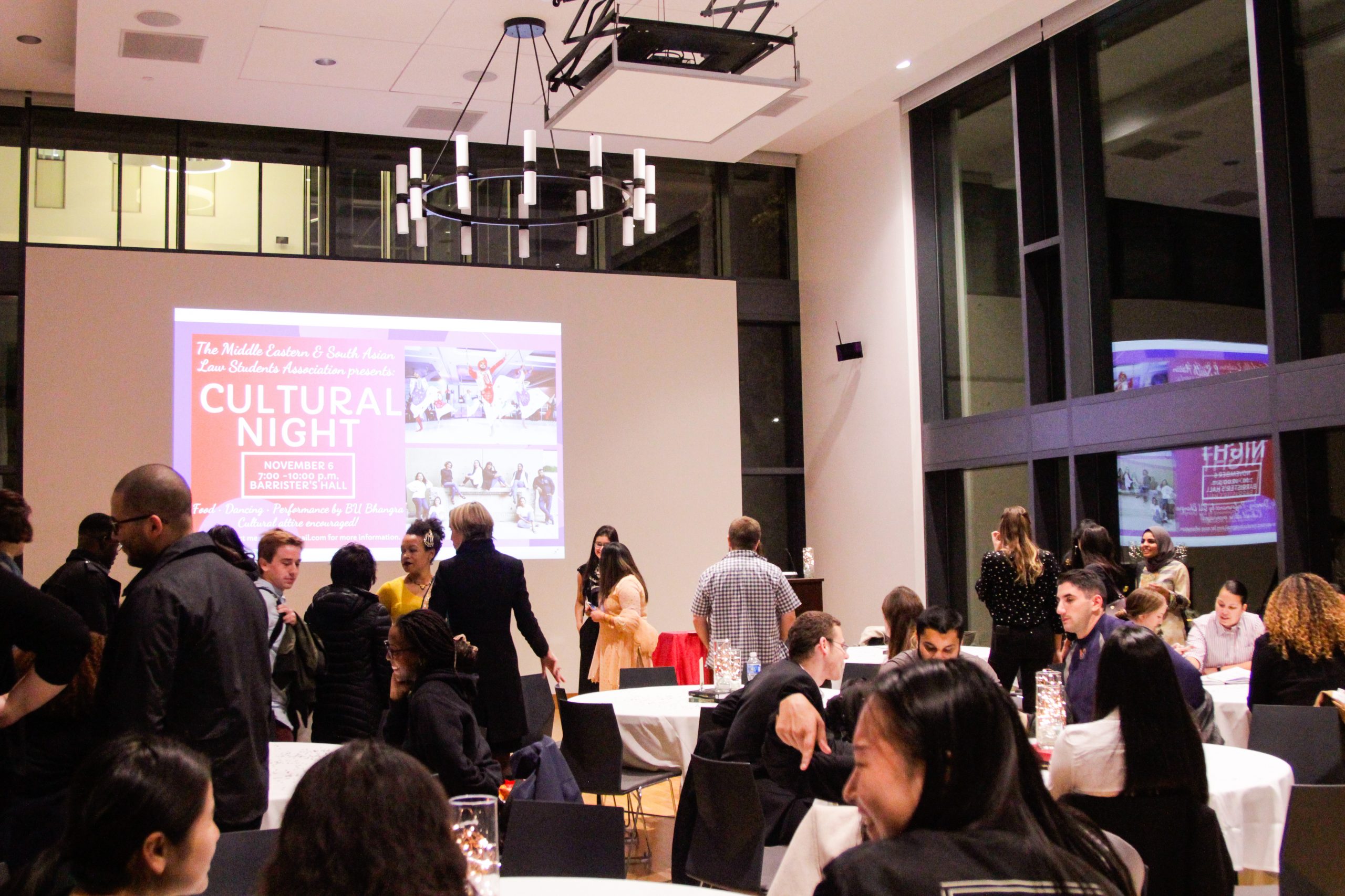 seated view of a full house at MESALSA Cultural night, students and professors talking in front of a projector