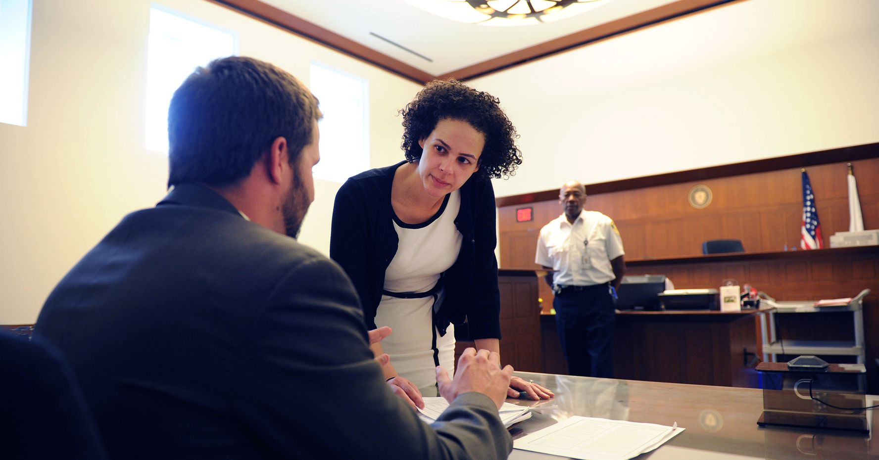 Two students discuss a case in a large courtroom. A bailiff stands in the background.