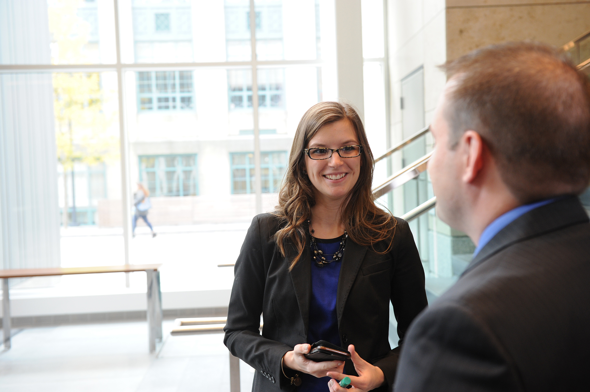 A student with long hair and glasses talks with a classmate in the atrium.