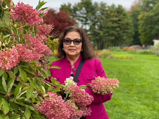 Nasim Parveen, wearing a fuchsia jacket, stands next to a large flowering plant. A green lawn and garden is in the background. 