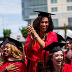 3 smiling BU graduates in red gowns and black mortarboard caps, with one standing and clapping.