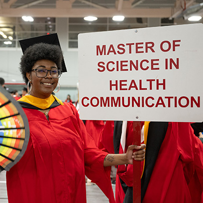 A graduate posing at the MET convocation ceremony, holding a sign that reads “Master of Science in Health Communication”