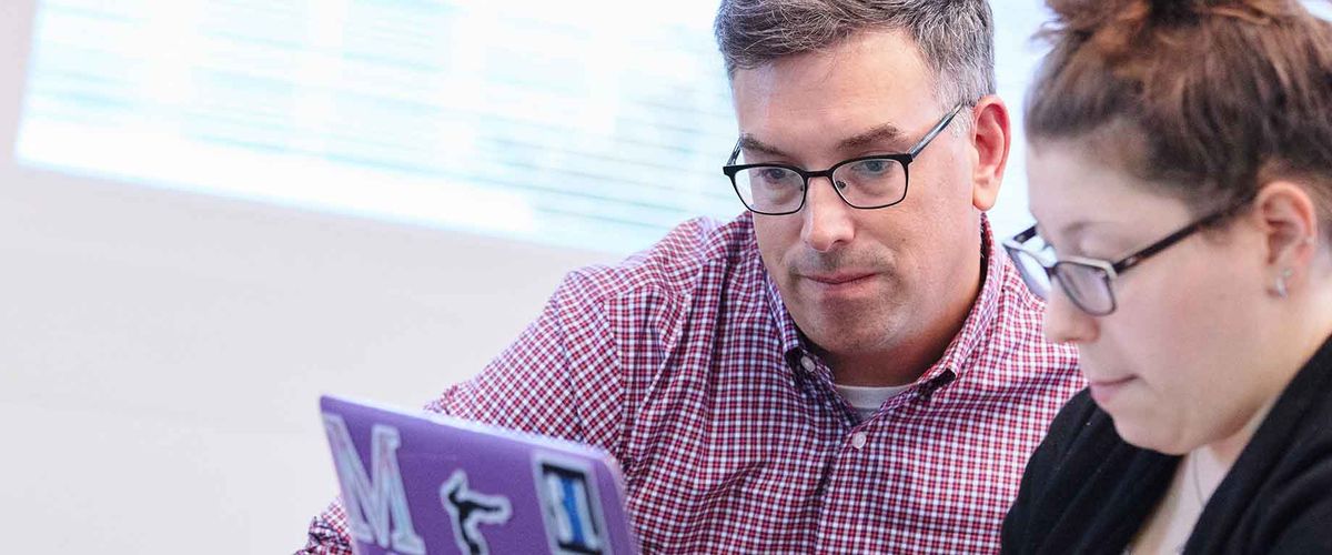 MET Professor Shea Cronin works with a student on a laptop in a classroom