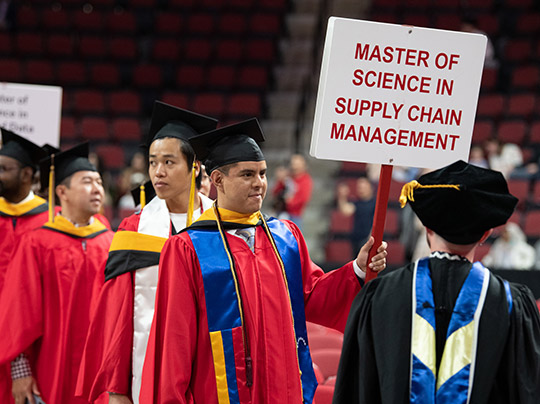 A graduate walking during the MET convocation ceremony, holding a sign that reads “Master of Science in Supply Chain Management”