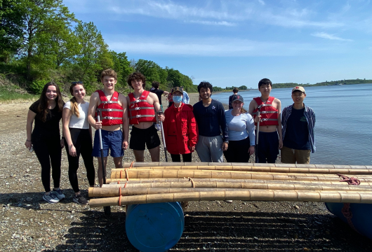 A group of undergrad honors students pose by the shore of a lake with life jackets on