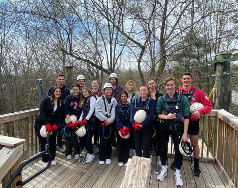 A group of over 10 smiling undergrad honors student stand on a zip line platform in the woods with harnesses and helmets.