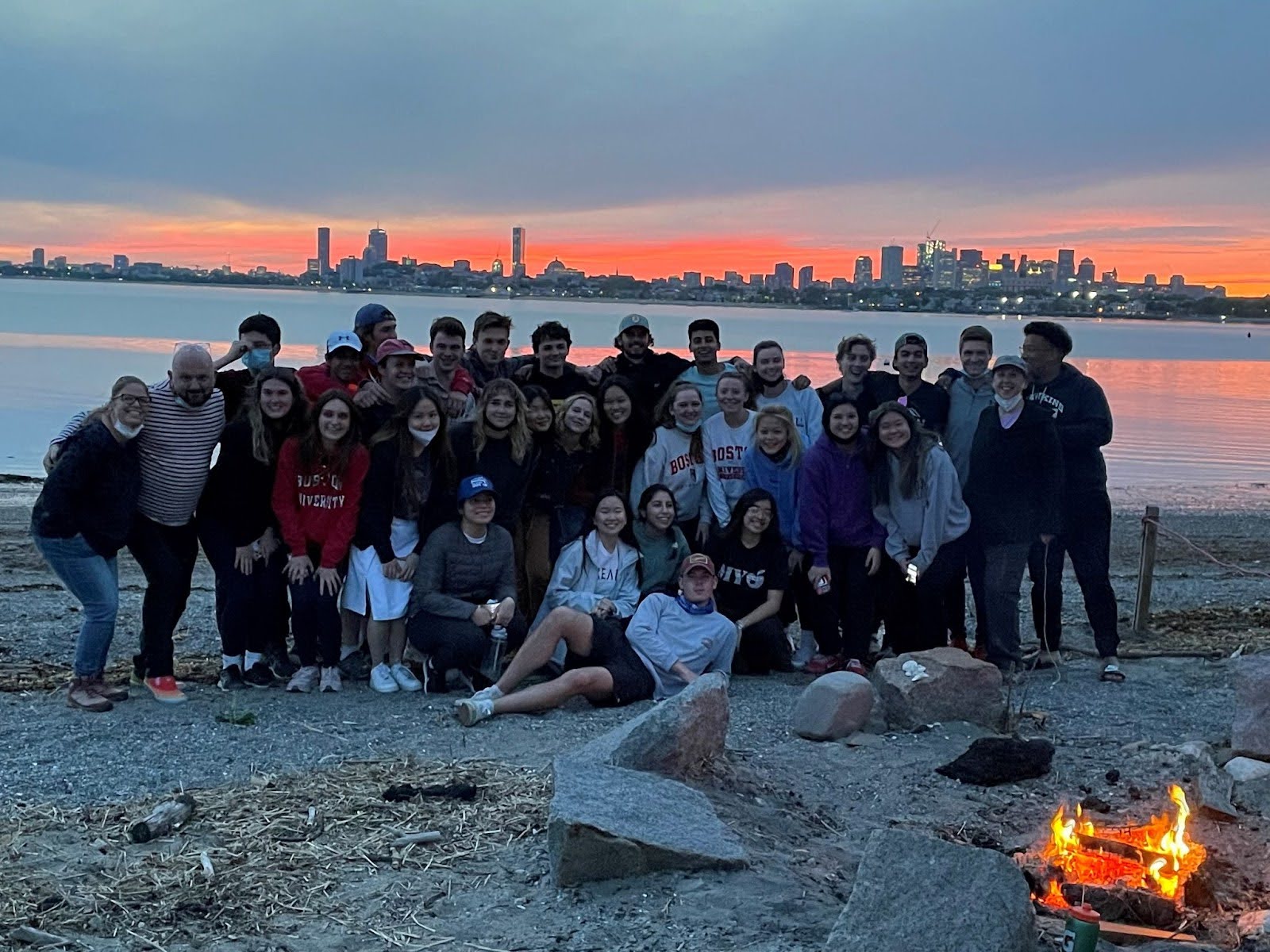 A group of undergrad honors student stand on the beach next to a campfire with the Boston skyline in the background at twilight.