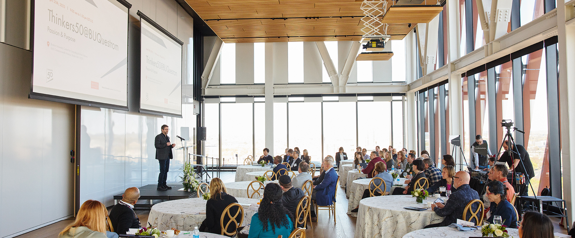 Man at the front of an event space speaking before a crowd gathered around round tables