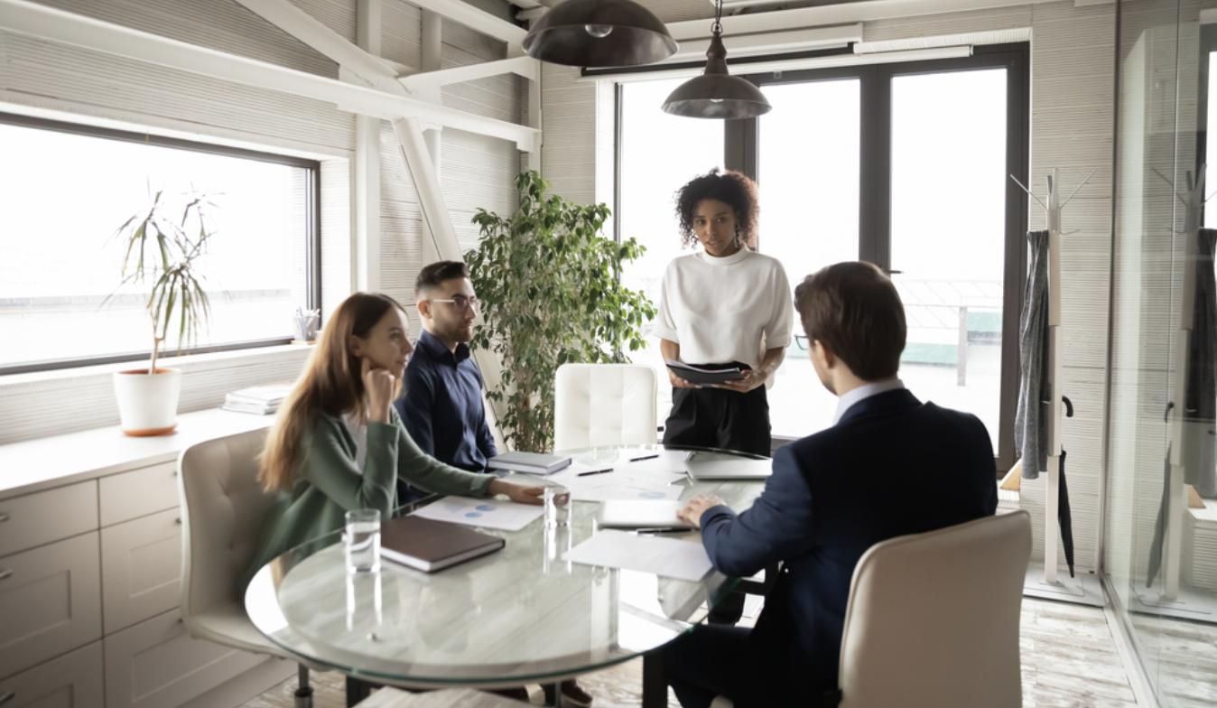 A businesswomen stands in front of colleagues at a conference table in a modern meeting room.
