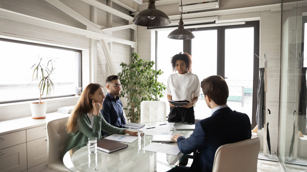 3 business people sit around a table in a modern office building while one person is standing leading the conversation.