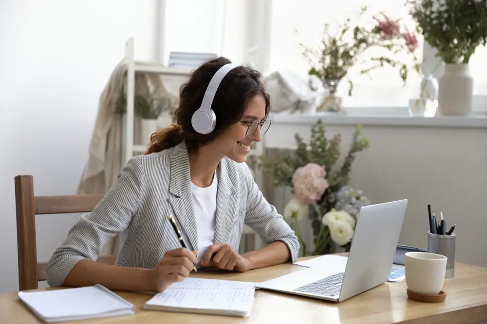 Young women wearing business casual clothing taking notes and smiling while interacting with someone on laptop.