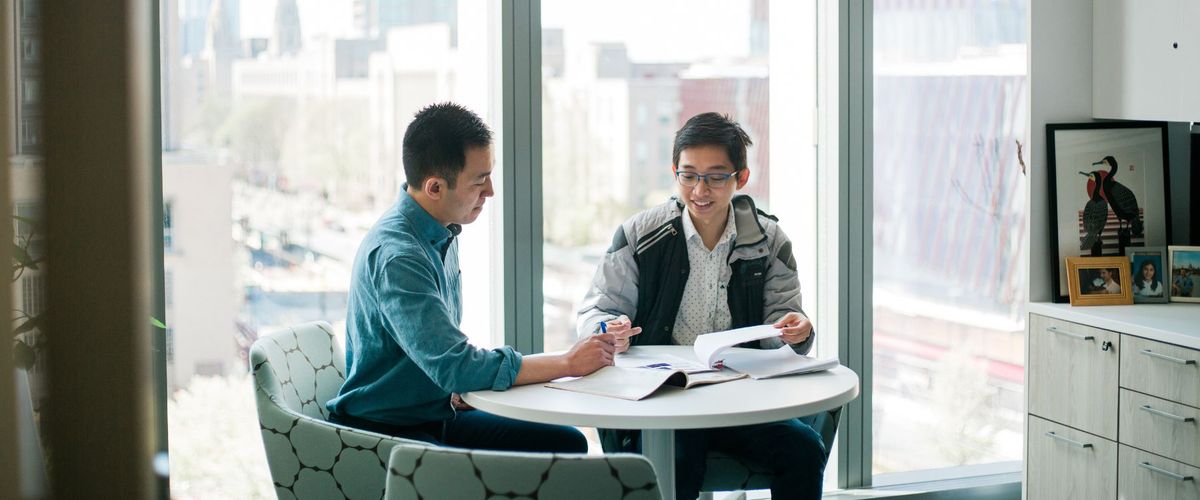 Two people sit at a table in front of a window. They are looking at papers together and having a lively discussion.