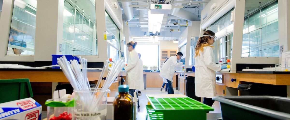 Several people stand at lab benches wearing white coats and protective eye gear. In the foreground, there are lab supplies.