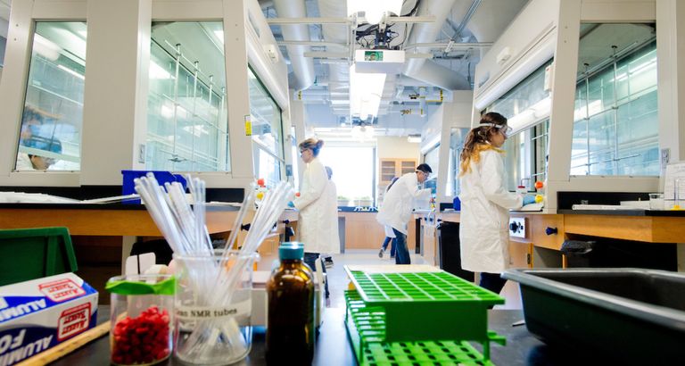Several people stand at lab benches wearing white coats and protective eye gear. In the foreground, there are lab supplies.