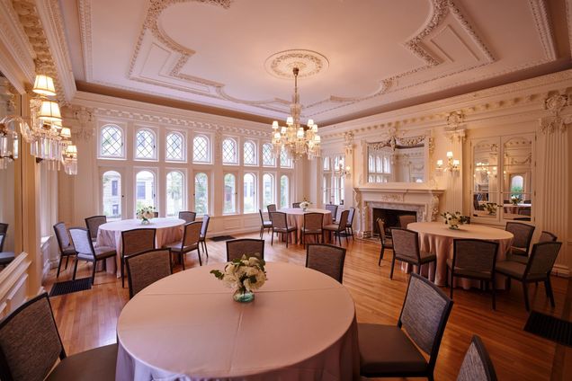 Round tables & chairs setup in the ivory-walled Music Room with chandeleir light turned on.