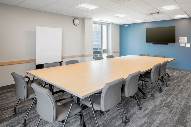Room view of empty conference table with chairs, TV mounted on a blue wall, moveable marker board, and large window.