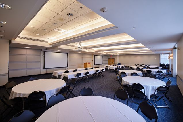 Angular view of Colloquium Room setup with round & rectangular tables with a BU podium and 2 screens for projection.