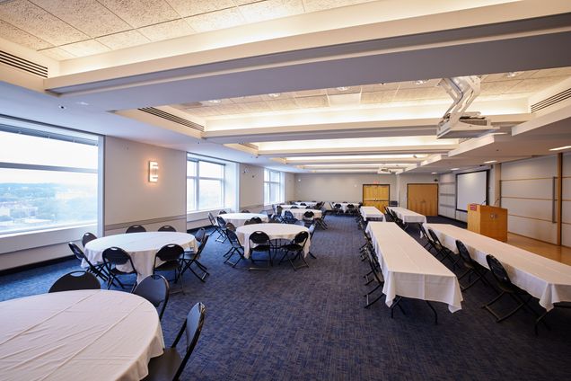 Side-long view of Colloquium Room setup with round & rectangular tables with a BU podium and 2 screens for projection.
