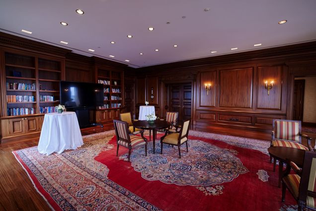 Empty view of Kenmore Room, set with 2 high-top tables with white tablecloths, a circular wood table with 4 yellow-upholstered chairs, on a red ornate carpet and wood paneled walls.