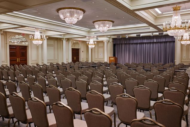 Empty Ballroom setup with grey chairs in rows, some with classroom tables, a podium with BU logo in the front next to a grey curtain.