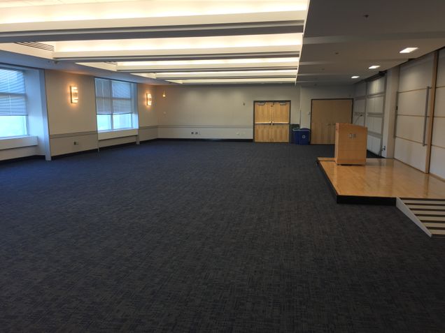 Empty room view of Colloquium Room with dark carpet, light walls, and low stage wooden platform with podium.