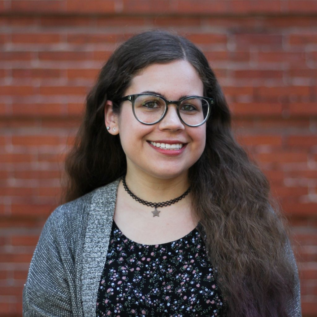 Peyton Veytia, with long wavy brown hair and glasses, wearing a colorfully speckled black top and grey sweater, stands against a brick backdrop, smiling at the camera