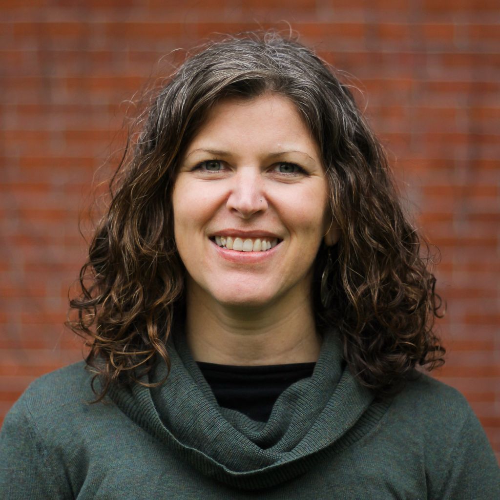 Cathleen Prata, with curly short brown hair, wearing a grey cowl-neck sweater and black top, stands in front of a brick backdrop, smiling at the camera