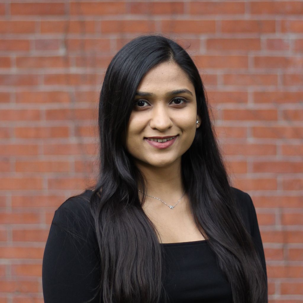 Kanish Mittal, with long, dark hair, wearing a long-sleeve black top, smiles in front of a brick backdrop.